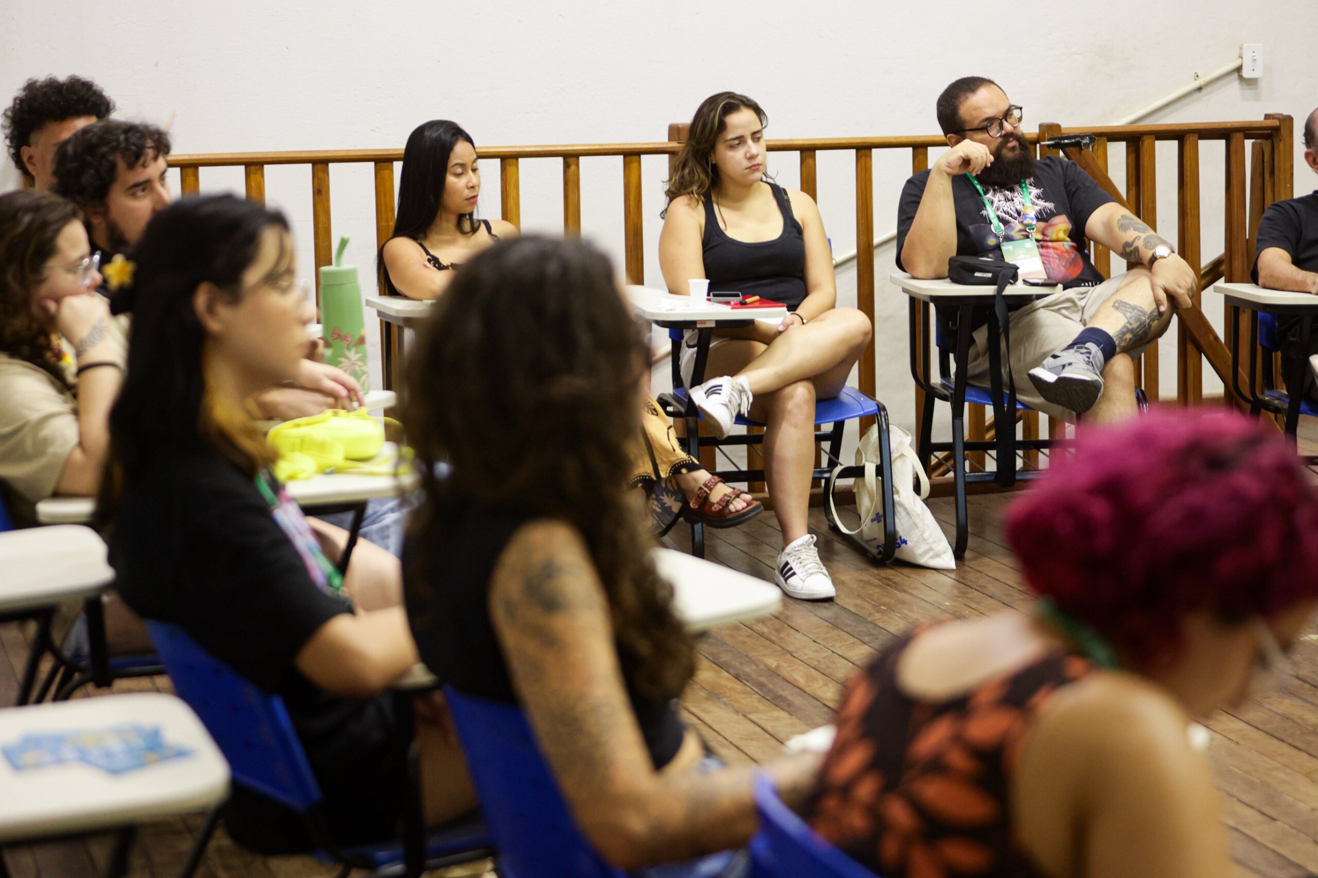 Fotografia de um grupo de pessoas sentadas em carteiras universitárias, em uma sala com piso de madeira e parede clara. As pessoas aparecem em círculo, ouvindo atentamente uma atividade formativa ou conversa coletiva. Algumas usam crachás e estão com materiais pessoais sobre as mesas, como garrafas, cadernos e celulares. A cena transmite um momento de escuta, participação e troca em ambiente acadêmico ou cultural.