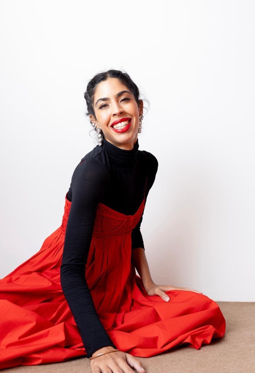 Mulher sentada no chão, sorrindo para a câmera, diante de um fundo branco. Ela usa uma blusa preta de manga longa por baixo de um vestido vermelho volumoso. O cabelo está preso, e ela usa batom vermelho e brincos. A pose é descontraída, com uma das mãos apoiada no chão e a outra sobre o vestido.