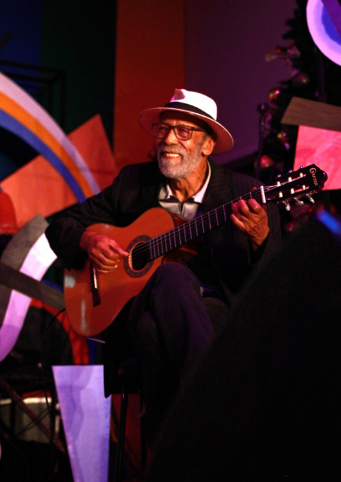 Fotografia de um homem idoso tocando violão em uma apresentação musical. Ele usa chapéu claro, óculos e roupa escura, e aparece sorrindo enquanto segura o instrumento. A cena ocorre em um ambiente de palco ou espaço cultural, com iluminação colorida em tons de vermelho, roxo e azul. Ao fundo, há elementos decorativos e parte de outro músico ou equipamento, mas o destaque principal é o artista tocando violão.