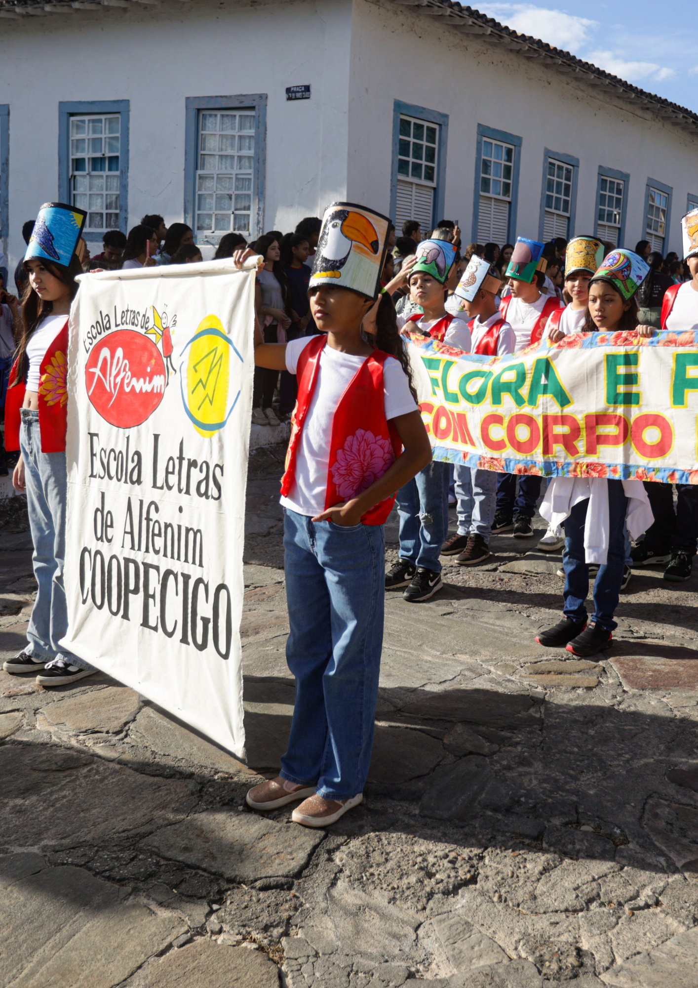 Fotografia de crianças participando de uma caminhada em uma rua histórica de pedra, diante de construções antigas com paredes brancas e janelas azuis. Algumas crianças seguram faixas e estandartes, incluindo um banner da Escola Letras de Alfenim, da COOPECIGO, e outra faixa com a frase parcialmente visível “Flora e fauna com corpo”. Muitas usam adereços coloridos na cabeça com ilustrações de animais e elementos da natureza. Ao fundo, há outras crianças, jovens e adultos acompanhando a atividade.