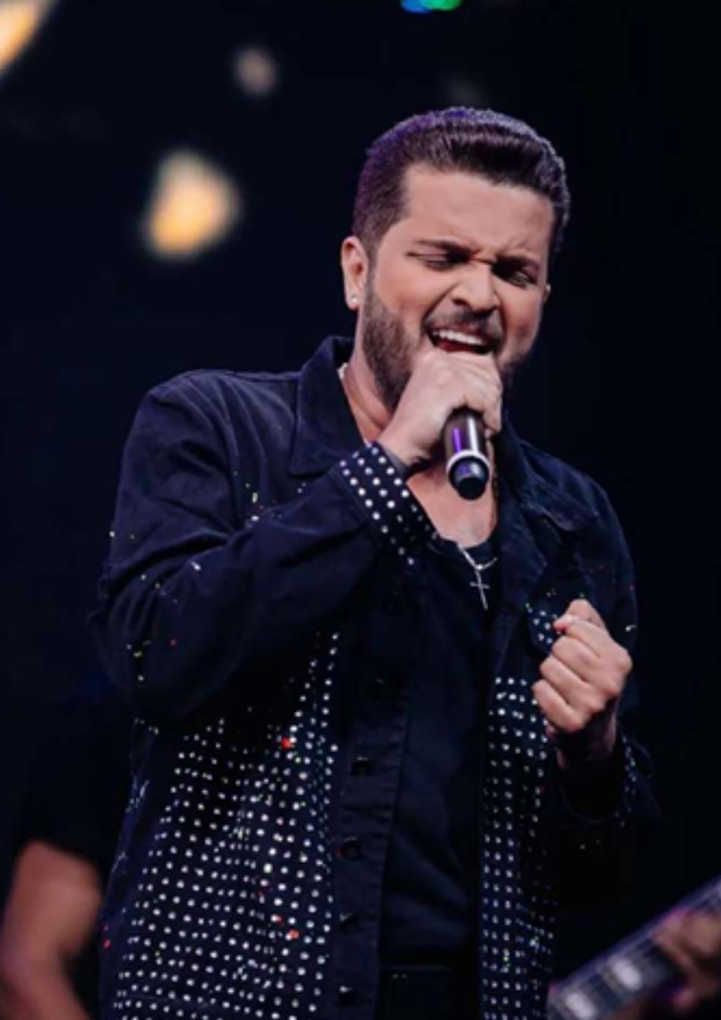 Fotografia de um cantor se apresentando no palco, segurando um microfone próximo à boca. Ele usa roupa escura com detalhes brilhantes e um colar com pingente em formato de cruz. A expressão facial indica intensidade durante a performance musical. Ao fundo, há iluminação de palco desfocada e parte de outro músico ou instrumento, reforçando o ambiente de show ao vivo.