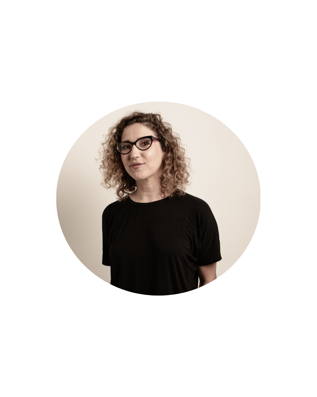 Retrato de uma mulher dentro de um recorte circular. Ela usa óculos, camiseta preta e tem cabelo cacheado claro. A pessoa aparece em frente a uma parede clara, olhando para a câmera com expressão leve.