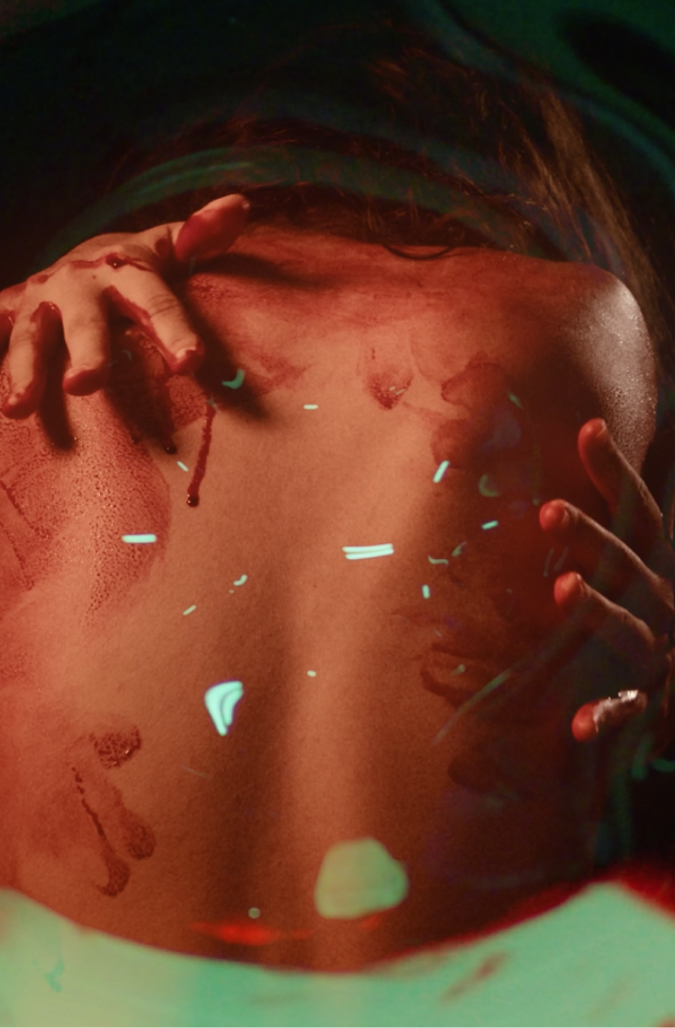 Fotografia em tons escuros de uma pessoa com o corpo parcialmente coberto por sombras e luzes coloridas. A imagem mostra as mãos e parte do tronco, com marcas vermelhas e reflexos luminosos em verde e vermelho sobre a pele.