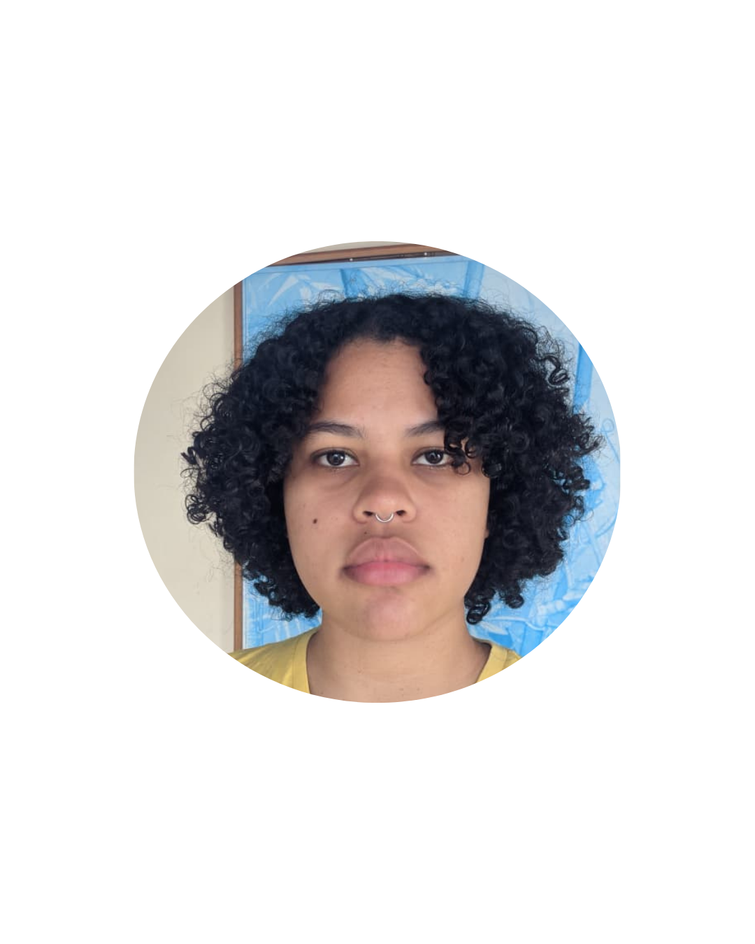 Retrato de uma pessoa dentro de um recorte circular. Ela tem cabelo curto e cacheado, usa camiseta amarela e piercing no nariz. A pessoa está olhando diretamente para a câmera, com fundo interno claro e um elemento azul parcialmente visível atrás.
