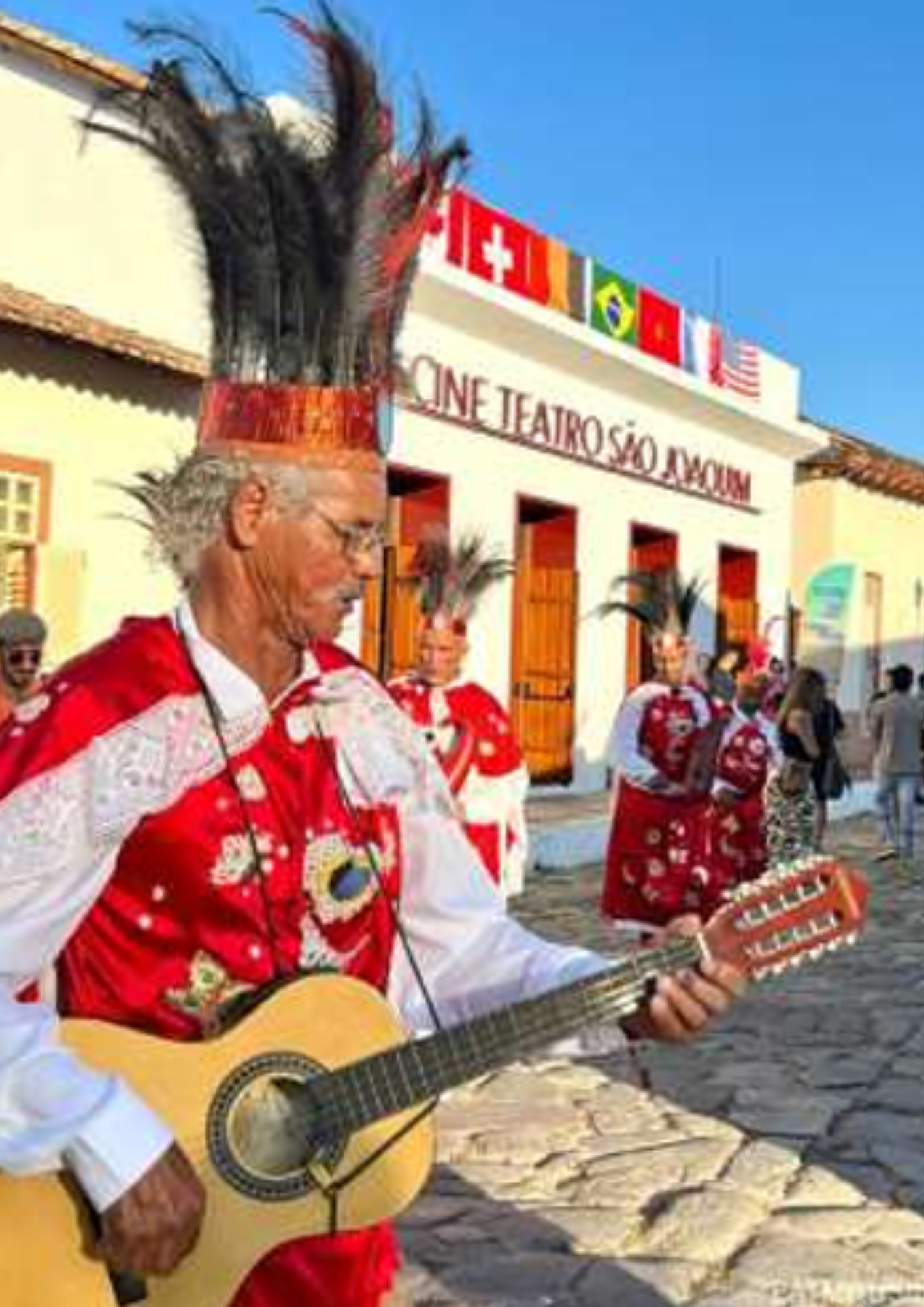 Homem tocando violão em uma rua de pedra, usando roupa vermelha e branca com estampas decorativas e um adereço alto de penas na cabeça. Ao fundo, outras pessoas usam trajes semelhantes e caminham próximas à fachada do Cine Teatro São Joaquim, onde há bandeiras de diferentes países na parte superior do prédio.