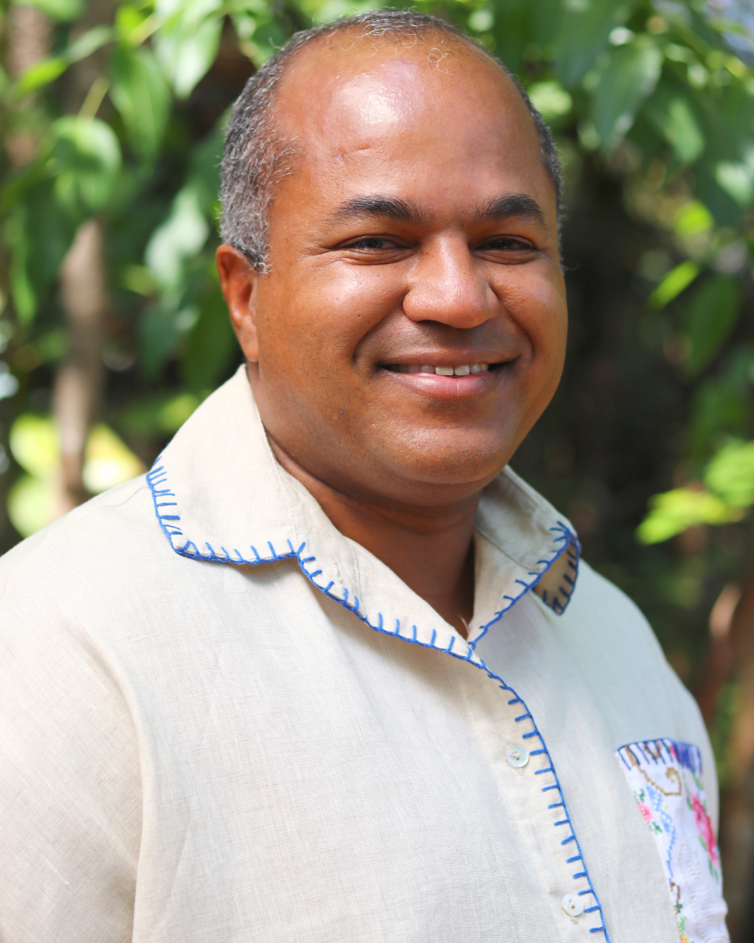 Fotografia em formato de retrato de um homem sorrindo, enquadrado do peito para cima. Ele usa uma camisa clara com detalhes bordados em azul na gola, na abertura frontal e no bolso. Ao fundo, há vegetação verde desfocada, indicando um ambiente externo com luz natural. A imagem destaca a pessoa em primeiro plano, transmitindo uma expressão acolhedora e tranquila.