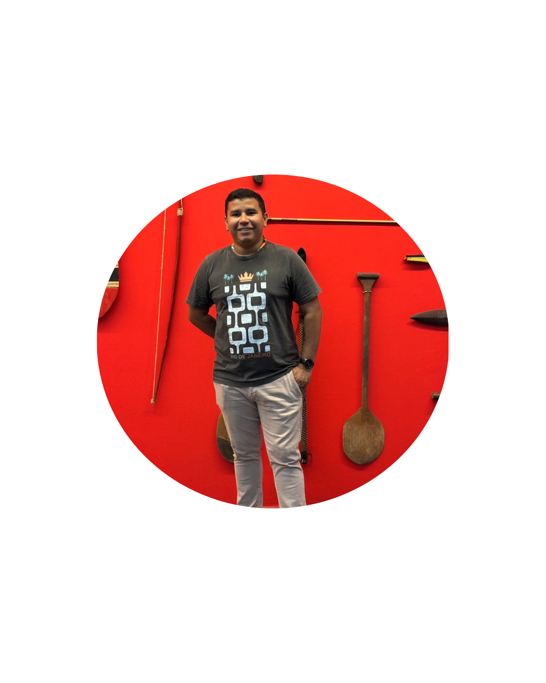 Retrato de um jovem dentro de um recorte circular. Ele está em pé diante de uma parede vermelha com objetos alongados pendurados ao fundo. Usa camiseta cinza, calça clara e relógio, com uma das mãos no bolso e a outra atrás do corpo.