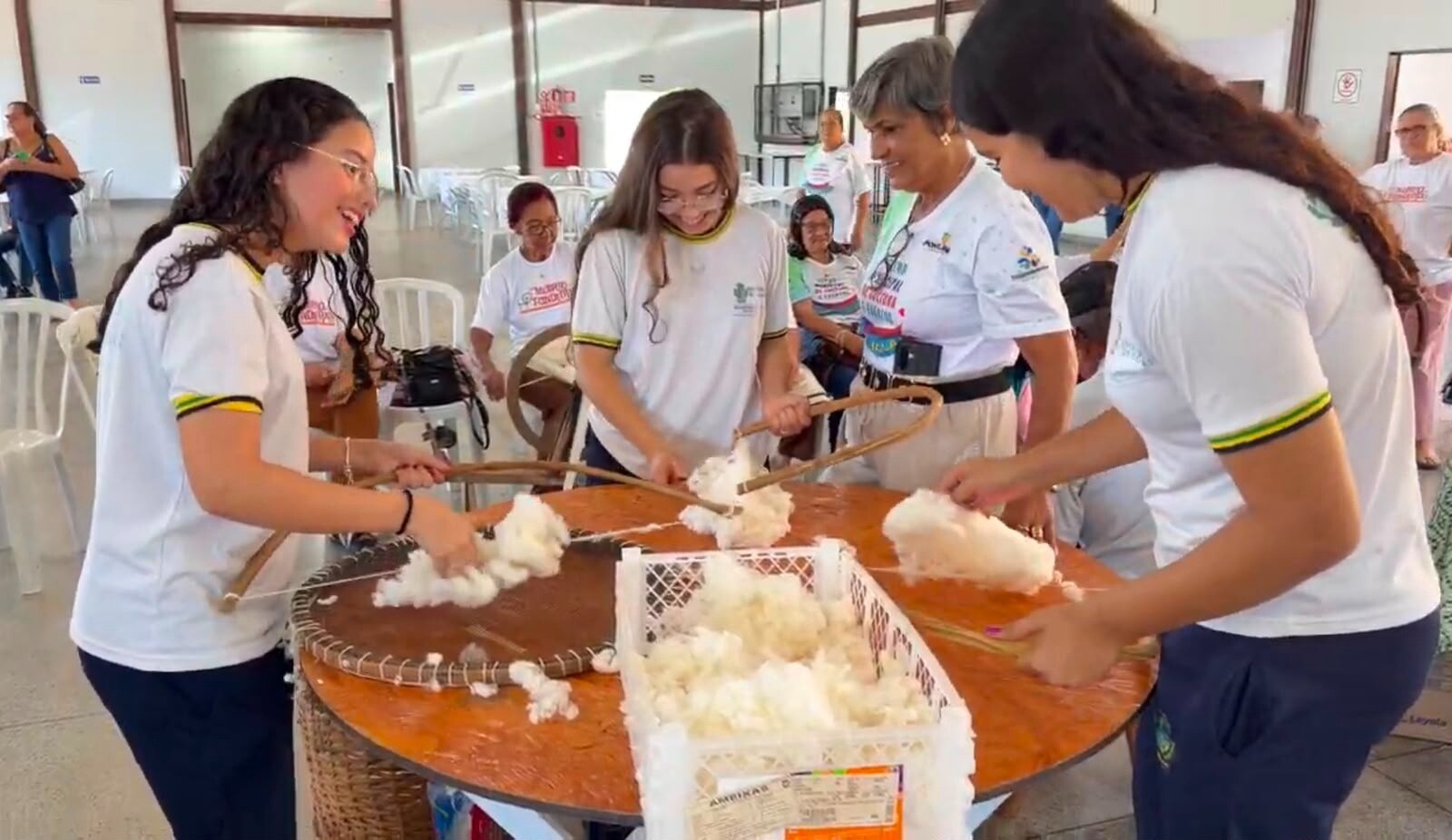 Grupo de pessoas reunido ao redor de uma mesa redonda em um salão amplo, realizando uma atividade manual com algodão. Três jovens em primeiro plano usam camisetas brancas e manuseiam arcos ou instrumentos de madeira sobre a mesa. Ao fundo, outras pessoas estão sentadas em cadeiras brancas, observando ou participando da atividade.