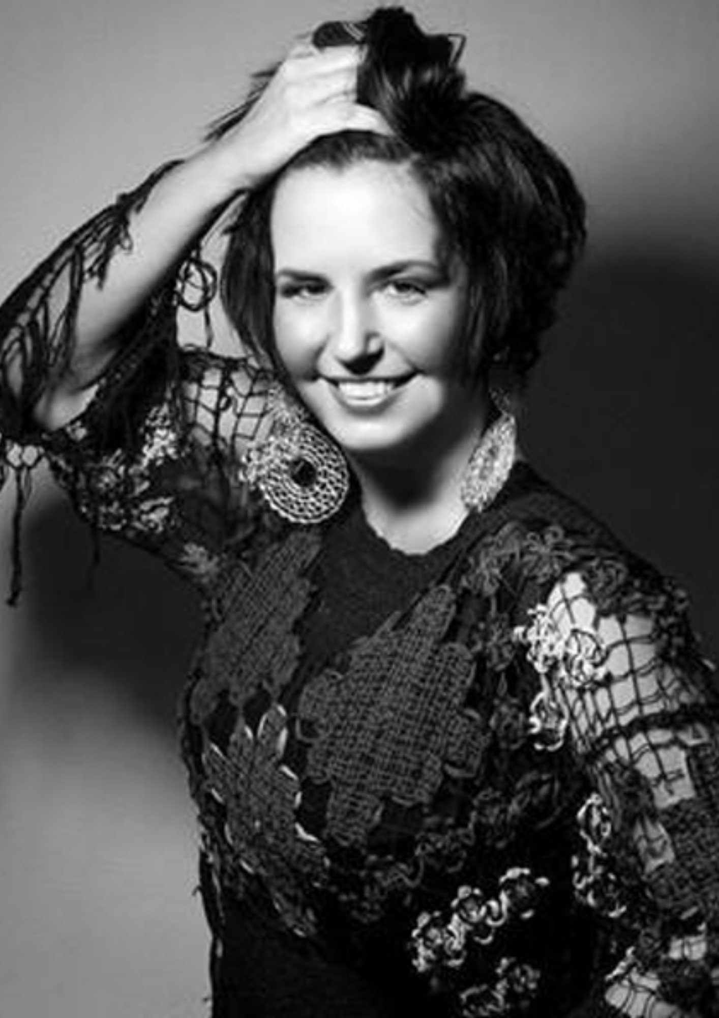 Fotografia em preto e branco de uma mulher sorrindo, em pose de retrato. Ela está com uma das mãos apoiada sobre a cabeça e usa uma roupa escura com transparências, renda e detalhes bordados. Também utiliza brincos grandes e circulares. O fundo é neutro e desfocado, destacando a pessoa em primeiro plano. A imagem transmite um tom artístico e expressivo.