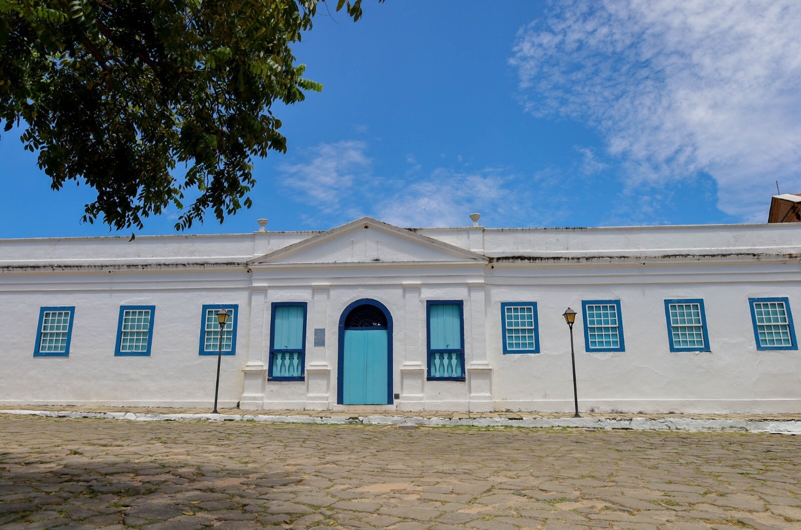 Fotografia de uma construção histórica branca com portas e janelas azuis, localizada diante de uma rua de pedra. A fachada possui uma porta central azul, janelas simétricas com molduras azuis e detalhes arquitetônicos em relevo. Há postes de iluminação em frente ao prédio, uma árvore à esquerda e céu azul com nuvens ao fundo.