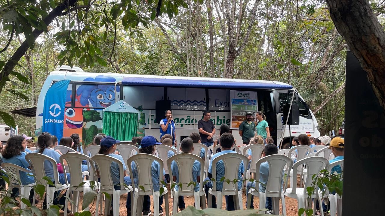Fotografia de uma atividade ao ar livre em uma área arborizada. Crianças estão sentadas em cadeiras brancas, voltadas para uma equipe que fala diante de um ônibus identificado com elementos visuais da Saneago e do Expresso Ambiental. Há banners, uma pequena estrutura com cortina verde e materiais informativos próximos ao veículo.
