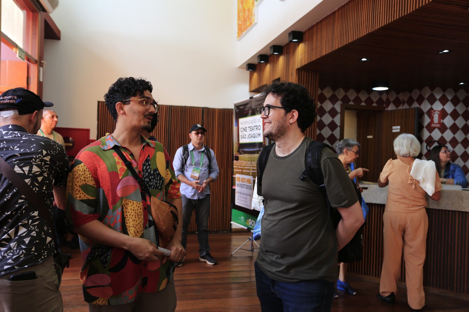 Fotografia de pessoas conversando no saguão de um espaço cultural. Em primeiro plano, dois homens conversam sorrindo; um usa camisa colorida estampada e o outro usa camiseta verde com mochila. Ao fundo, há outras pessoas circulando, balcão de atendimento, paredes com revestimento de madeira e um banner do Cine Teatro São Joaquim.