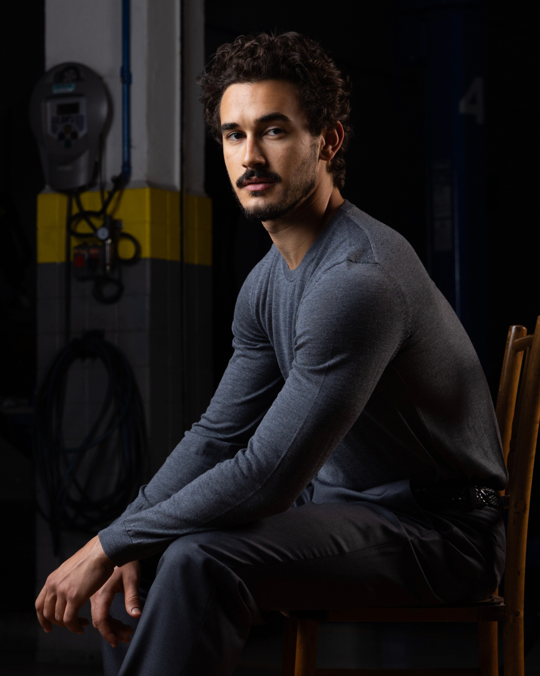 Homem adulto com cabelos escuros e cacheados, barba e bigode aparados, sentado de perfil em uma cadeira de madeira e olhando em direção à câmera. Veste camiseta de manga longa cinza e calça escura. A iluminação destaca seu rosto e parte superior do corpo, criando contraste com o ambiente ao fundo, que é escuro e lembra um espaço industrial ou técnico, com equipamentos, cabos e estruturas visíveis de forma desfocada. A expressão é séria e contemplativa, transmitindo um tom sóbrio e introspectivo.