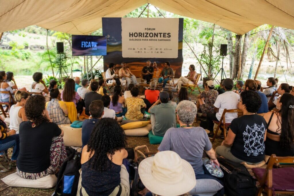 Registro de um painel ou roda de conversa realizado sob uma tenda, em ambiente externo e arborizado. Ao centro, há pessoas sentadas em cadeiras participando da discussão, com microfones e estrutura de som. O público acompanha a atividade sentado em cadeiras, almofadas e bancos, disposto em semicírculo. Ao fundo, aparece um painel com a identificação “Fórum Horizontes — Diálogos para novos caminhos” e uma tela lateral com a marca do FICA 2026.