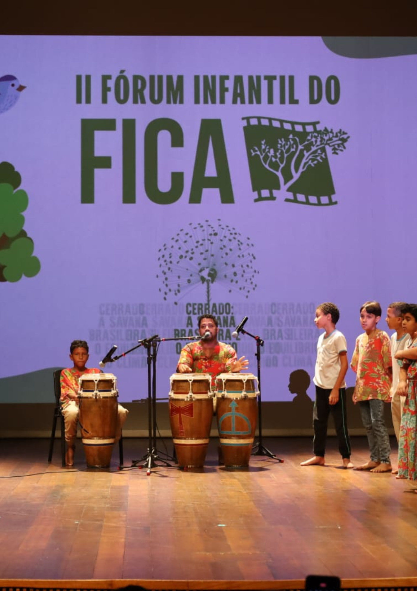 Fotografia de uma apresentação infantil em palco durante o II Fórum Infantil do FICA. Ao fundo, há uma projeção com o texto “II Fórum Infantil do FICA” e ilustrações relacionadas ao Cerrado. No centro do palco, um adulto toca tambores e conversa ao microfone, enquanto uma criança sentada também está diante de um tambor. À direita, outras crianças acompanham a atividade em pé, algumas usando roupas coloridas. A imagem registra um momento de participação, musicalidade e formação cultural voltada ao público infantil.