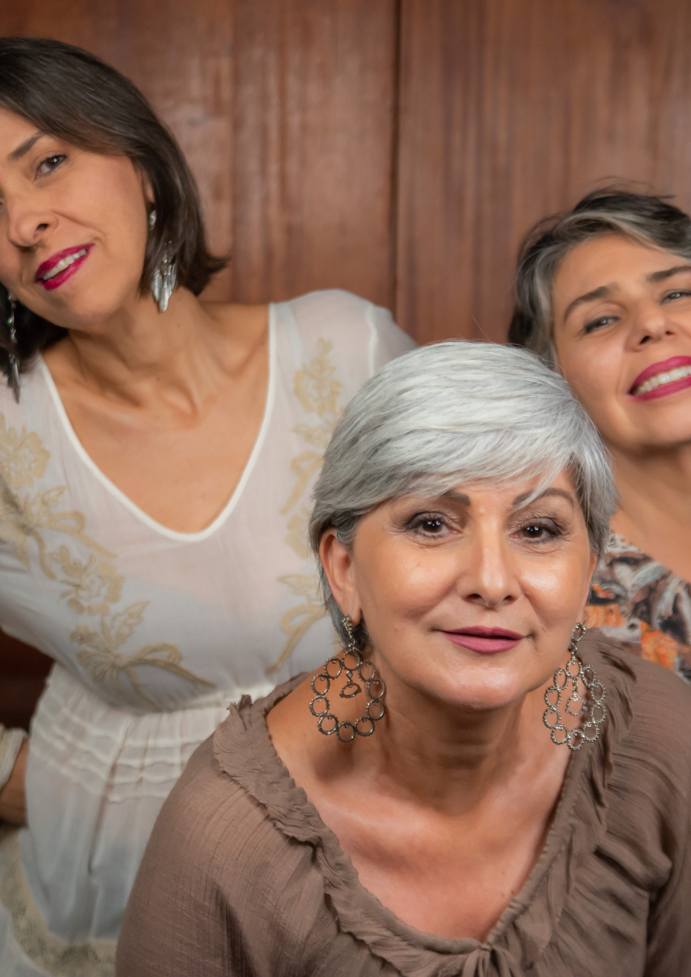 Três mulheres posando próximas à câmera, em frente a um fundo de madeira. A mulher ao centro está em primeiro plano, tem cabelo curto grisalho, usa blusa marrom e brincos grandes. À esquerda, aparece parcialmente uma mulher com cabelo escuro curto, usando roupa branca com detalhes bordados e brincos. À direita, outra mulher aparece parcialmente, sorrindo, com cabelo curto com mechas claras e batom rosa.