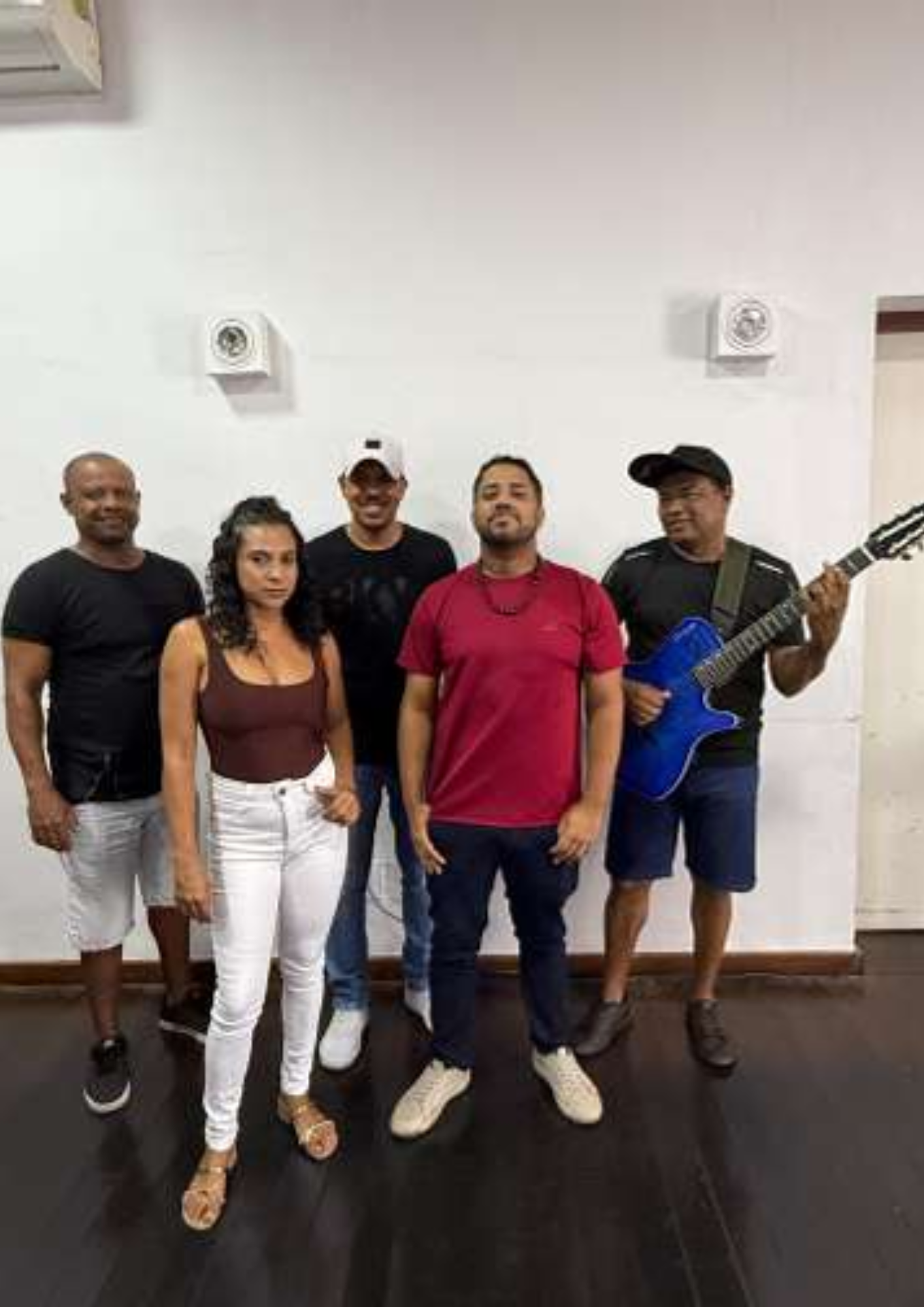 Fotografia de um grupo musical com cinco integrantes posando em uma sala de piso escuro e parede clara. Quatro homens e uma mulher aparecem em pé, voltados para a câmera. Um dos integrantes, à direita, segura um violão azul. A imagem sugere o registro de um grupo de samba ou apresentação musical, destacando os músicos reunidos em ambiente interno.