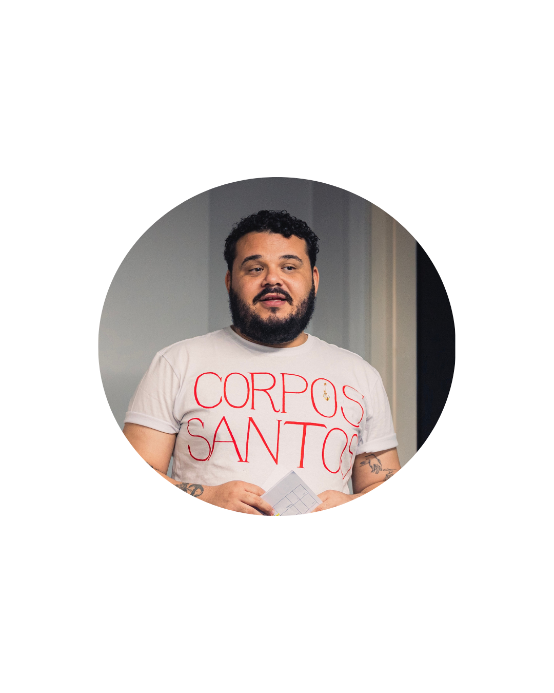Retrato de um homem dentro de um recorte circular. Ele usa camiseta branca com letras vermelhas, tem barba e segura algumas folhas nas mãos. Ao fundo, aparece um ambiente interno com parede clara e parte de uma abertura escura.
