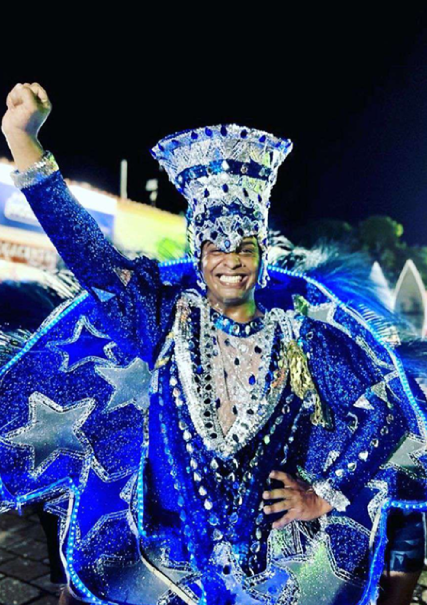 Fotografia de uma pessoa sorrindo durante uma apresentação ou desfile cultural, usando uma fantasia azul brilhante com detalhes prateados, estrelas e luzes decorativas. A pessoa aparece com um dos braços levantado e a outra mão na cintura, em pose expressiva e celebrativa. O traje inclui um adereço alto na cabeça e elementos volumosos nas costas, criando destaque visual. O fundo está escuro e desfocado, sugerindo um ambiente noturno de festa, espetáculo ou carnaval.