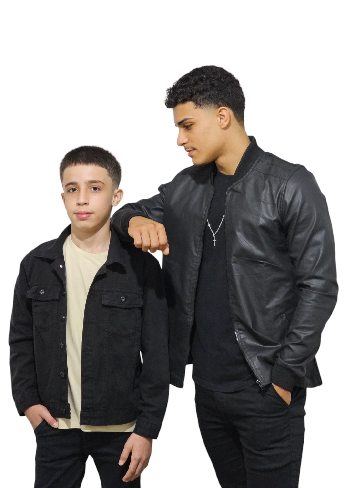 Dois jovens posando em frente a um fundo branco. À esquerda, um menino usa camiseta clara, jaqueta preta e calça preta, olhando para a câmera. À direita, um jovem mais alto usa camiseta preta, jaqueta preta de couro, calça preta e colar com pingente de cruz. Ele está com uma das mãos no bolso e o outro braço apoiado sobre o ombro do menino, olhando para ele.
