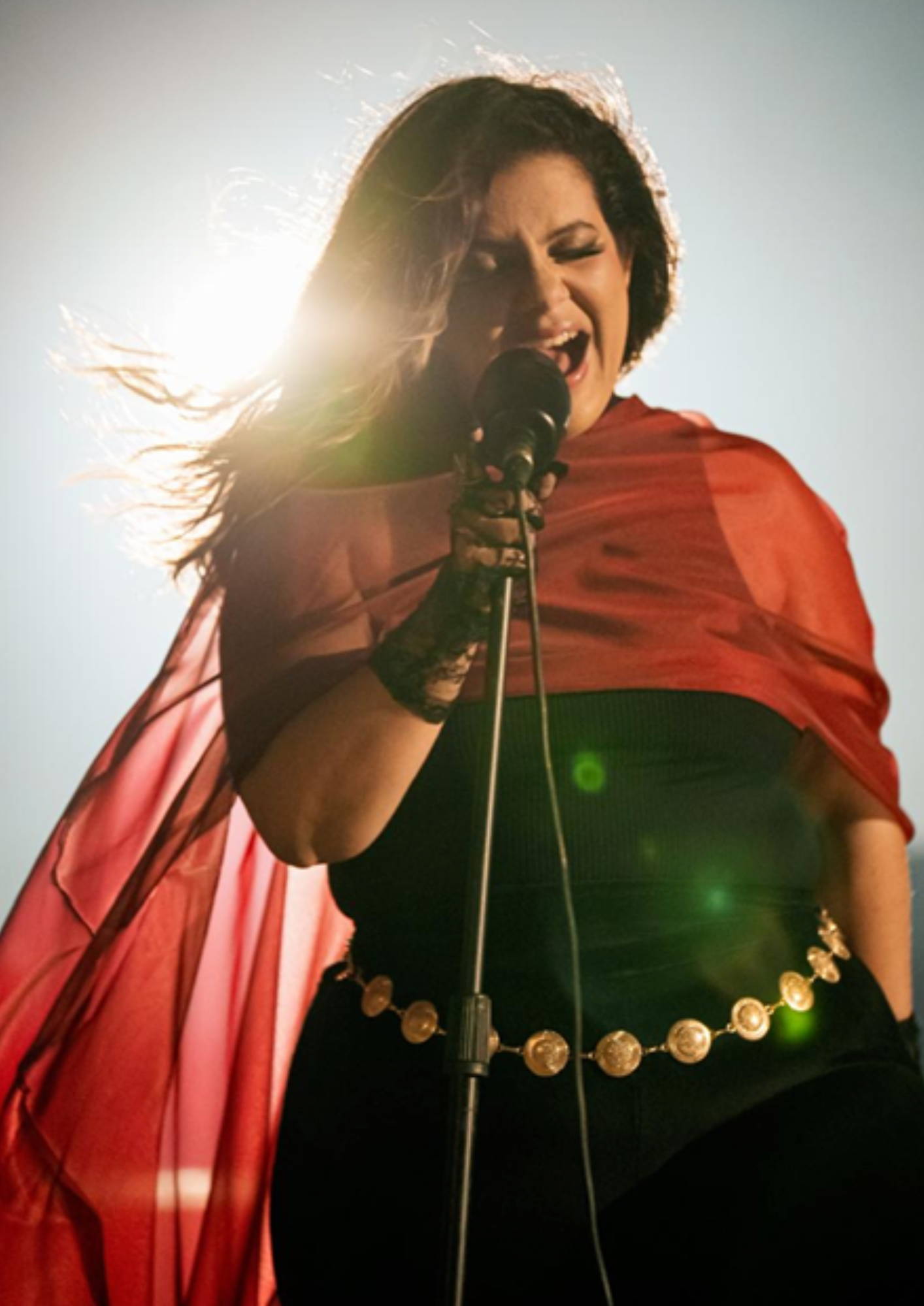 Cantora se apresentando ao ar livre, registrada em um enquadramento de baixo para cima, segurando um microfone enquanto canta com intensidade. Ela veste roupa preta acompanhada de uma capa ou tecido vermelho translúcido que se movimenta ao vento, criando um efeito dramático e elegante. A luz do sol ao fundo forma um forte contraluz, iluminando os contornos do cabelo e da vestimenta, além de produzir reflexos de lente na imagem. A artista aparece em destaque no centro da composição, transmitindo energia, emoção e presença de palco durante a apresentação.
