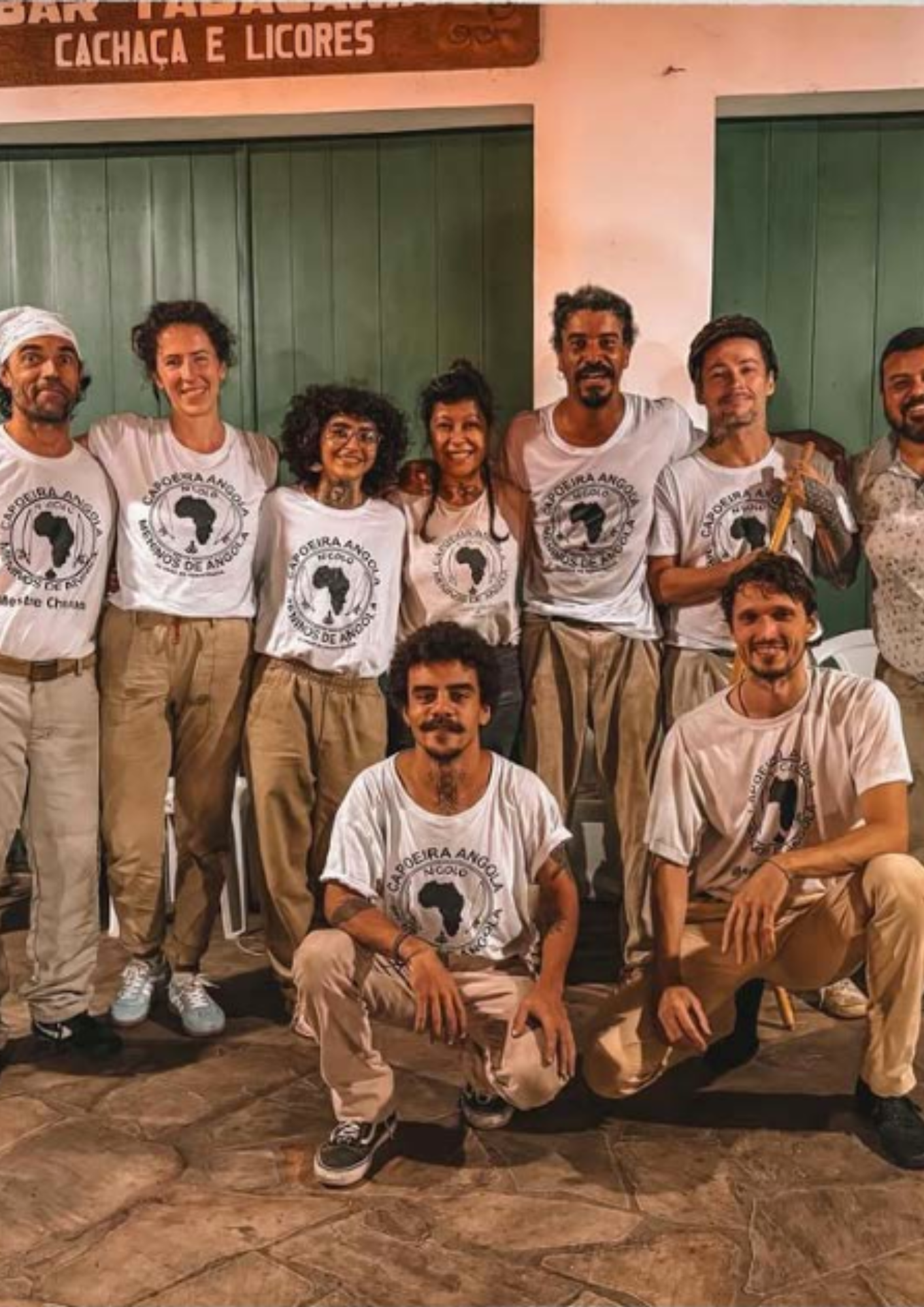 Grupo de nove pessoas posando em frente a uma fachada com portas verdes e parede clara. A maioria veste camiseta branca com estampa circular e calça em tons claros. Algumas pessoas estão em pé, lado a lado, enquanto duas estão agachadas à frente. Ao fundo, na parte superior, aparece parcialmente uma placa com os dizeres “Cachaça e Licores”.