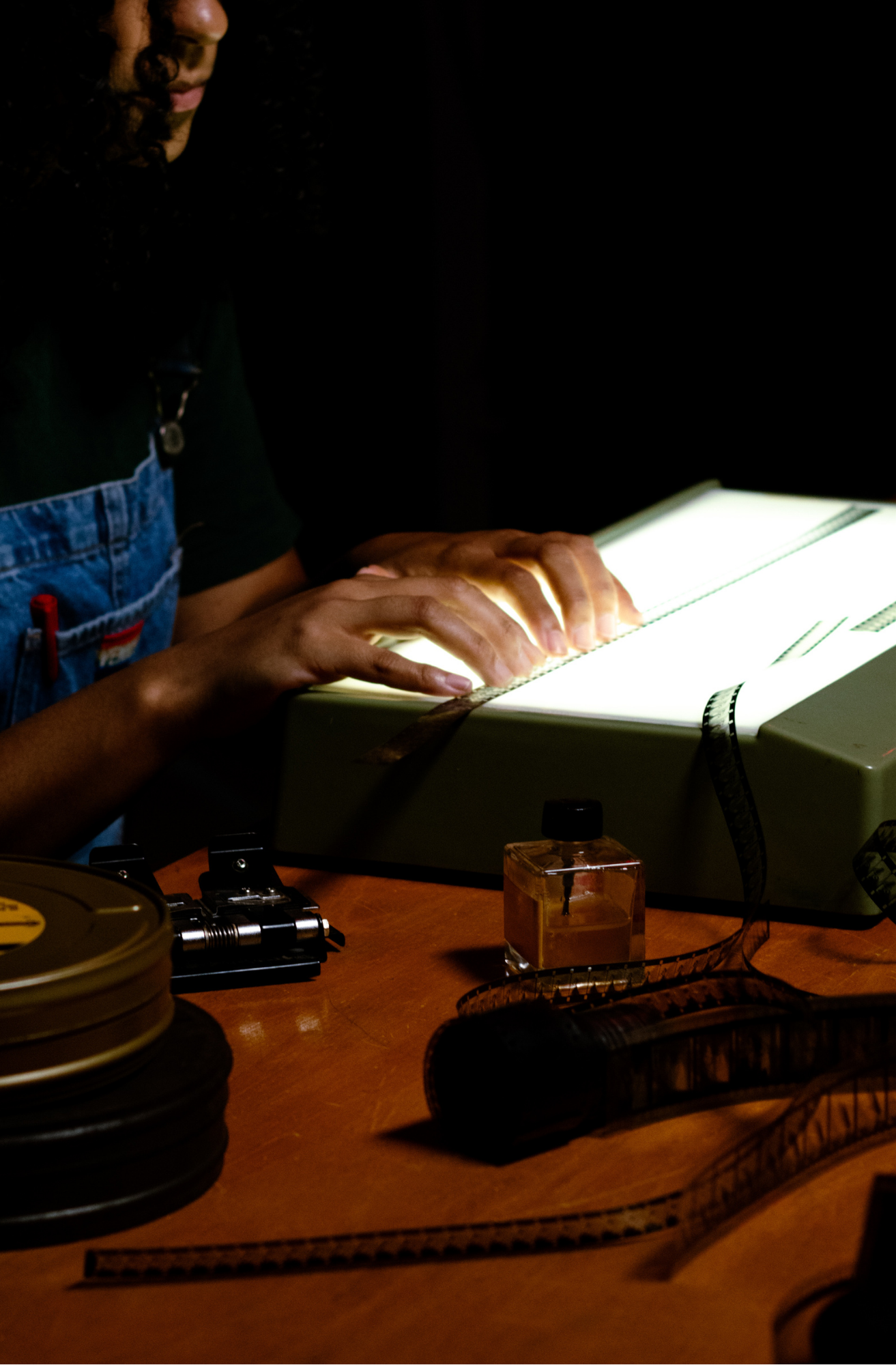 Fotografia de uma pessoa em ambiente escuro, manuseando tiras de filme sobre uma mesa iluminada. Há rolos de película, equipamentos e um frasco pequeno sobre a mesa. A iluminação destaca as mãos e o material cinematográfico.