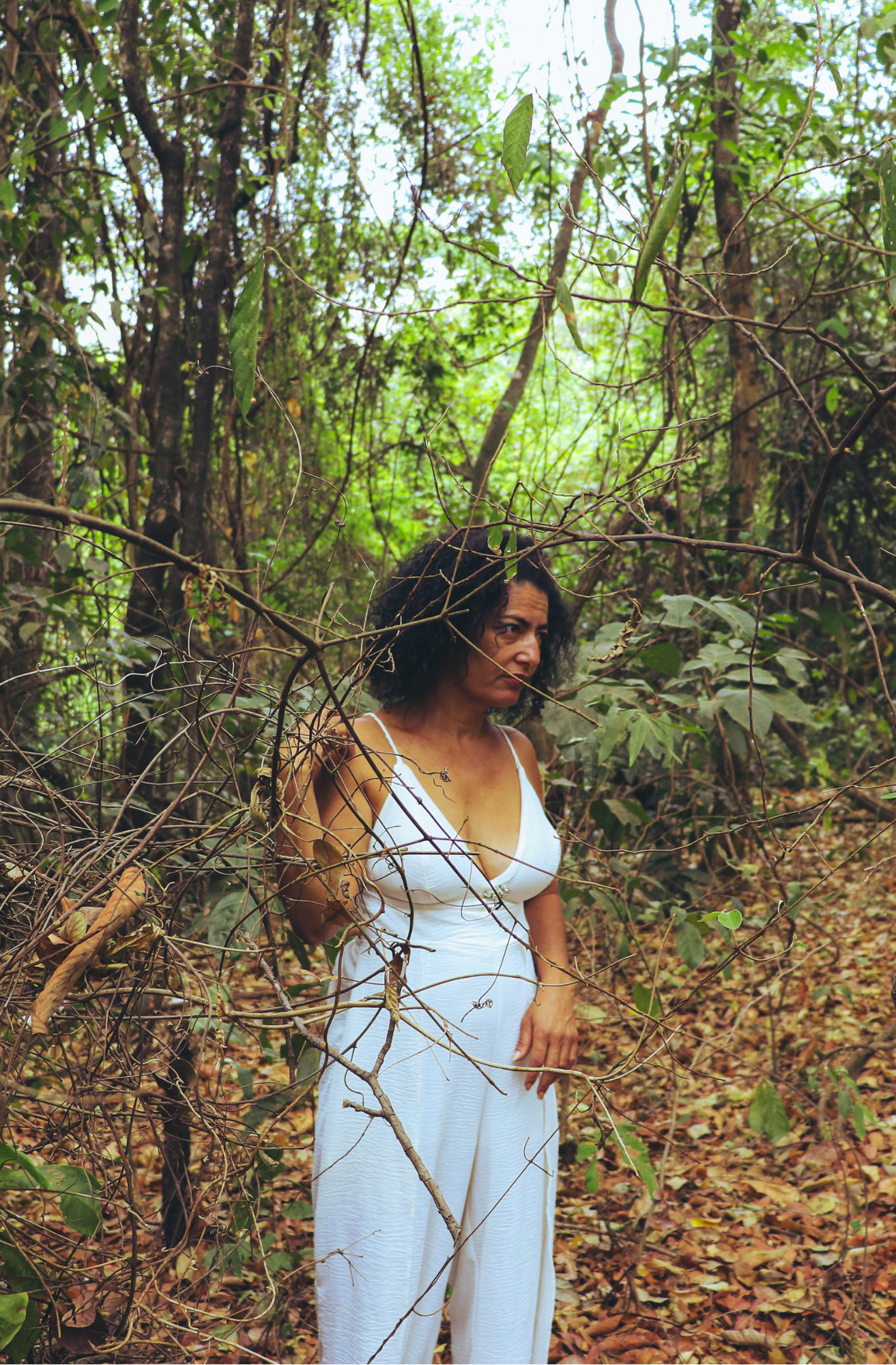 Fotografia de uma mulher em uma área de mata, usando roupa branca. Ela aparece entre galhos secos e vegetação densa, olhando para o lado. O cenário tem folhas no chão, árvores e luz natural filtrada pela vegetação.