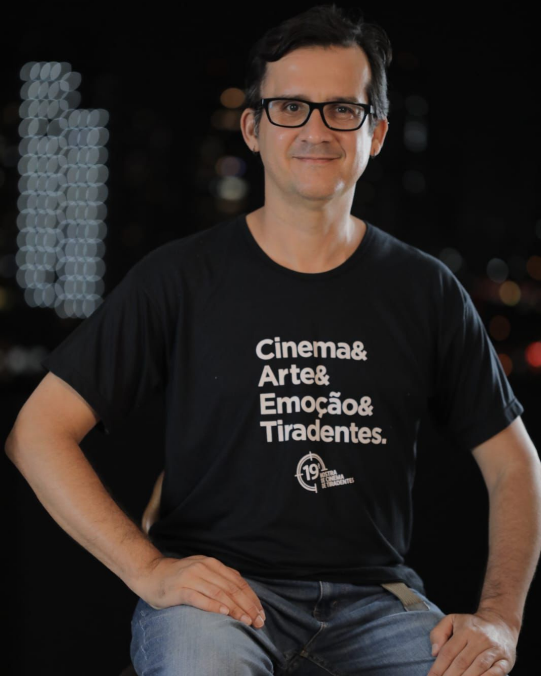 Fotografia de um homem sentado, usando óculos, camiseta preta com a frase “Cinema & Arte & Emoção & Tiradentes” e calça jeans. Ele aparece de frente para a câmera, com as mãos apoiadas nas pernas e uma leve expressão de sorriso. O fundo está escuro e desfocado, com pontos de luz ao fundo, sugerindo um ambiente noturno. A imagem tem aspecto de retrato e destaca a pessoa em primeiro plano.