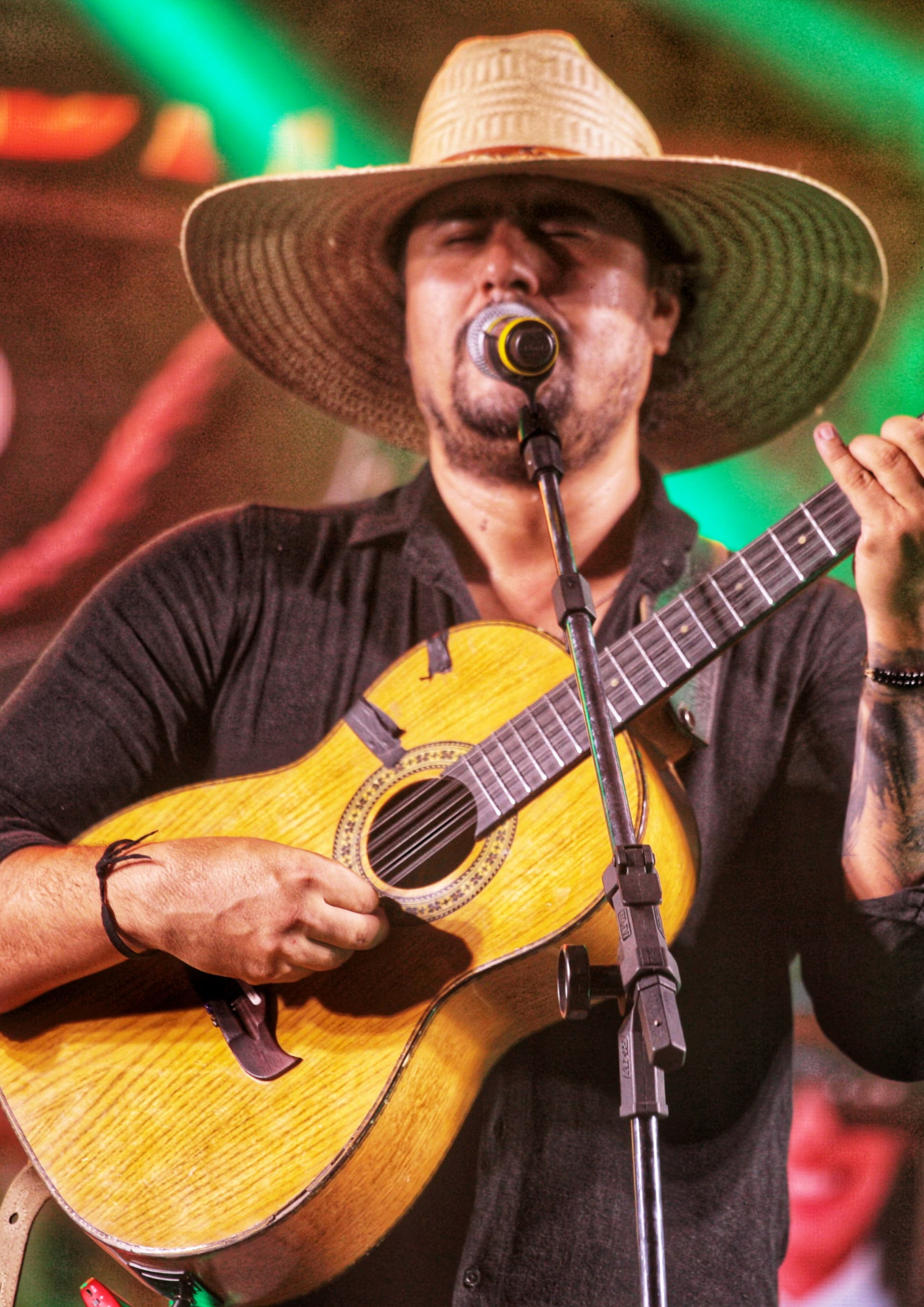Fotografia de um músico se apresentando no palco, cantando ao microfone enquanto toca violão. Ele usa camisa preta e um chapéu de palha de aba larga. A imagem destaca o artista em primeiro plano, com o violão ocupando boa parte da cena. Ao fundo, há luzes coloridas de palco em tons de verde e vermelho, criando um ambiente de apresentação musical ao vivo.