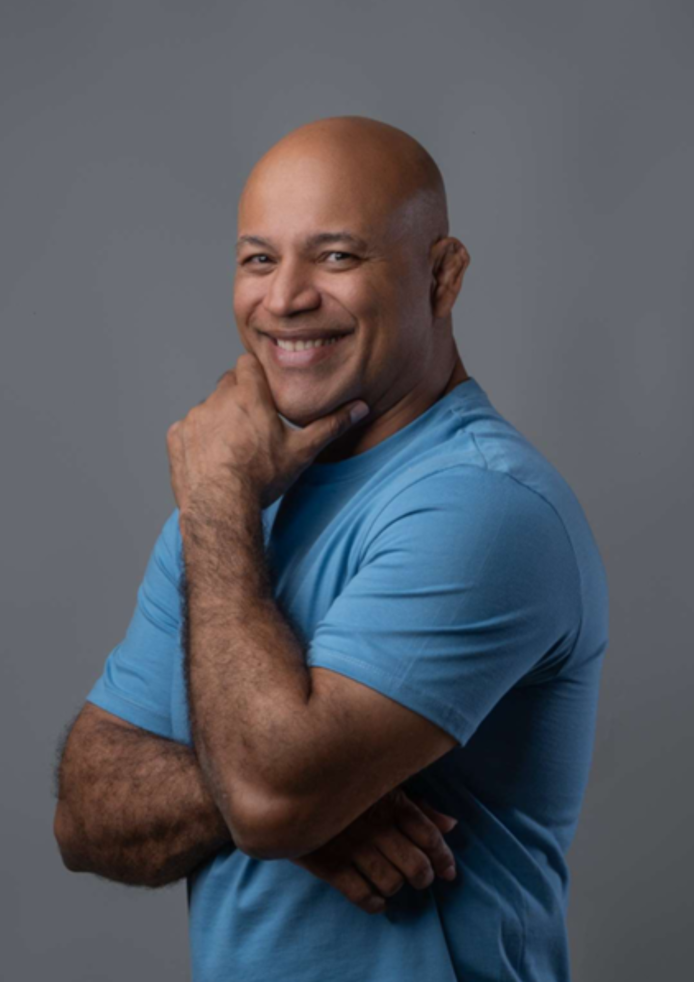 Fotografia em formato de retrato de um homem sorrindo, com os braços cruzados e uma das mãos apoiada no queixo. Ele usa camiseta azul e aparece de lado, voltado para a câmera. O fundo é cinza e neutro, com iluminação de estúdio, destacando a pessoa em primeiro plano. A imagem transmite uma expressão simpática, confiante e acolhedora.
