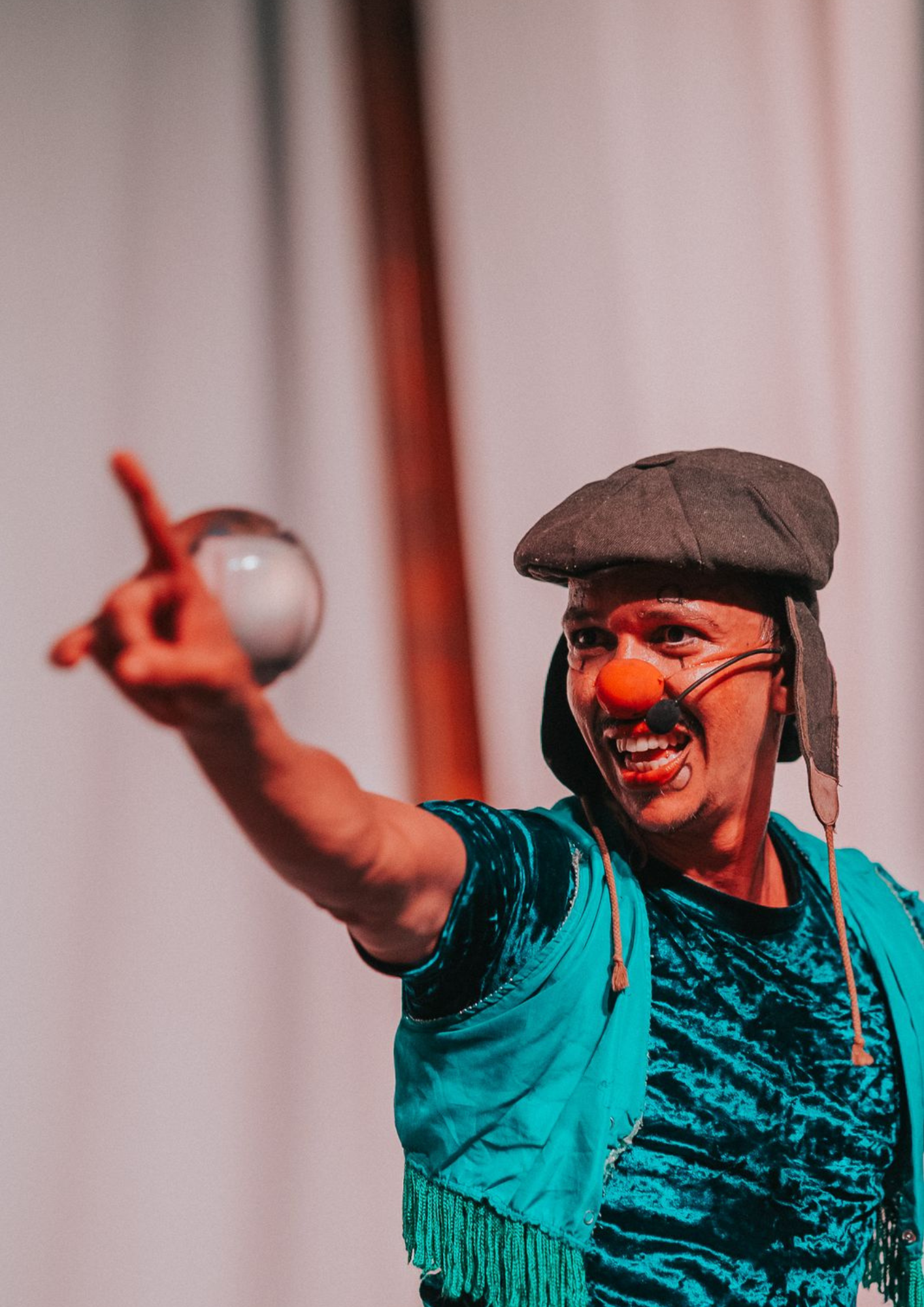 Pessoa em cena teatral ou circense, usando nariz vermelho, microfone de rosto, boina escura e roupa em tons de verde. Ela está com um dos braços estendido para frente, apontando com o dedo indicador, e segura um objeto circular desfocado na mão. A expressão facial é intensa, com a boca aberta, como se estivesse falando ou interpretando. O fundo é claro e desfocado, com iluminação avermelhada sobre a pessoa.