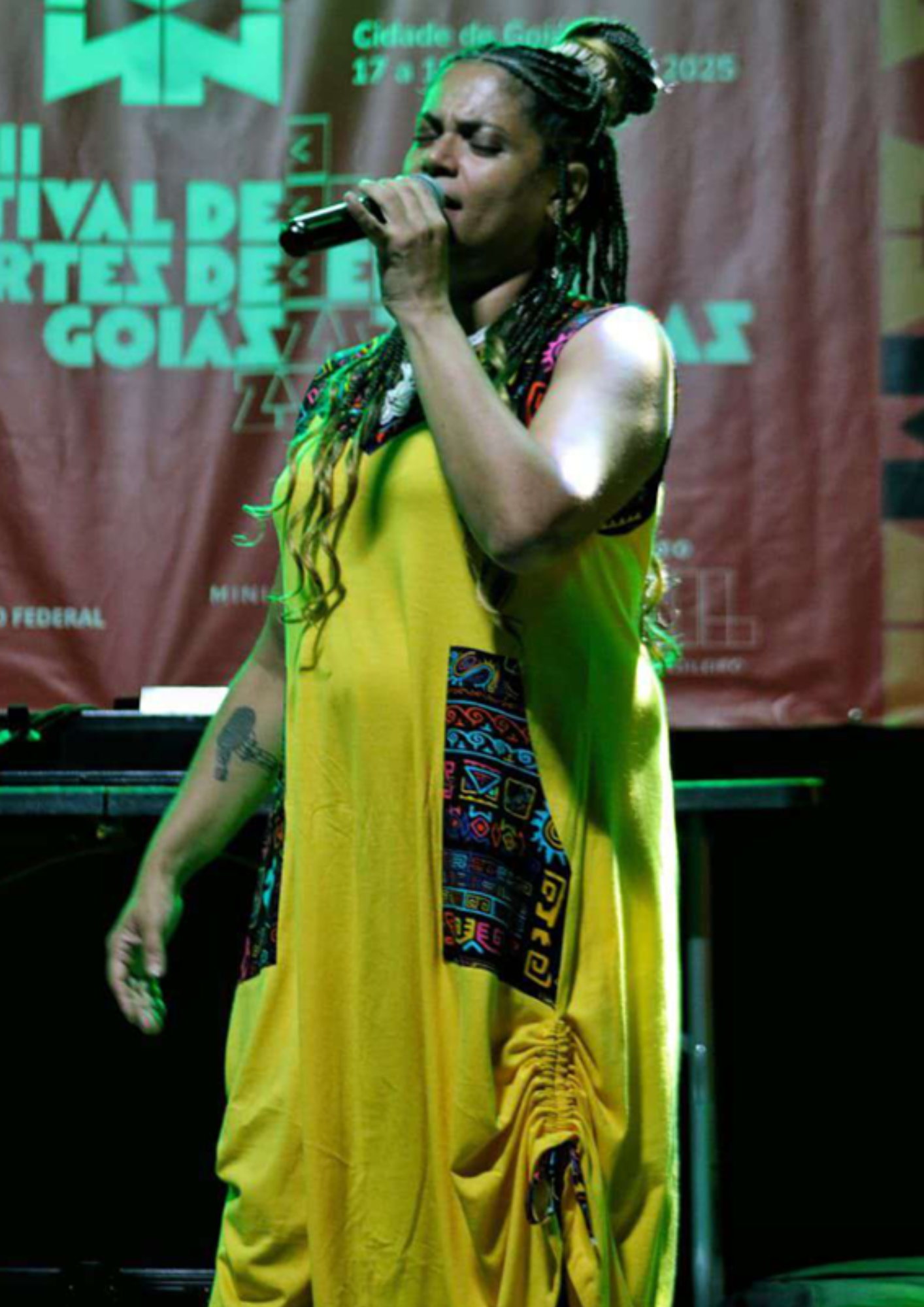 Pessoa cantando em um palco, segurando um microfone próximo à boca. Ela usa uma roupa longa amarela com detalhes coloridos nas laterais e tem o cabelo preso com tranças. A iluminação do palco tem tons de verde, e ao fundo aparece parcialmente um painel com textos relacionados a um festival de artes.