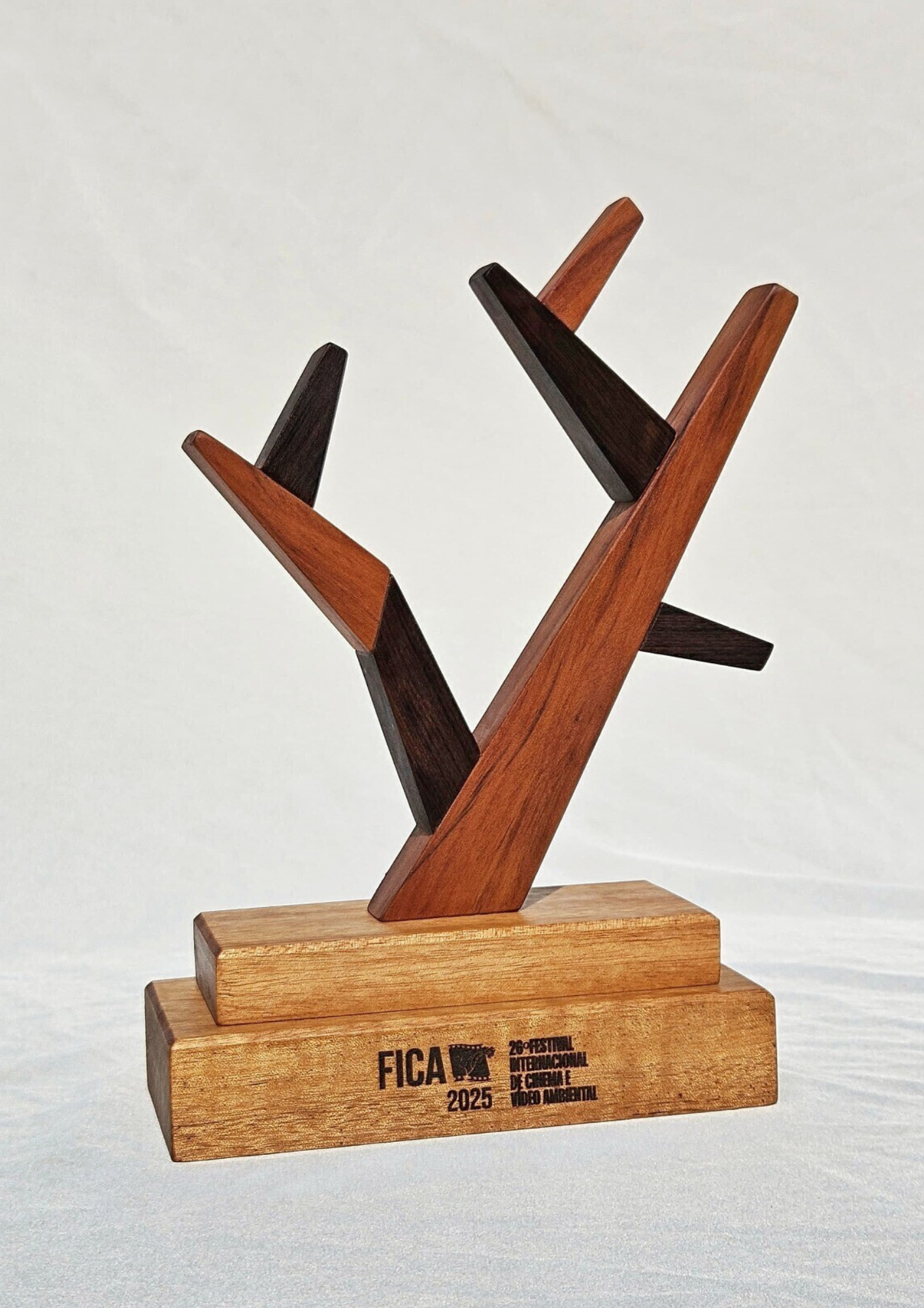 Troféu de madeira sobre fundo claro. A peça tem uma base retangular em dois níveis, com a marca “FICA 2025” e o texto “26º Festival Internacional de Cinema e Vídeo Ambiental” gravados na parte frontal. Acima da base, há uma estrutura formada por hastes de madeira em tons claros e escuros, dispostas em ângulos diagonais, criando uma composição abstrata e vertical.