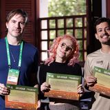 Fotografia de três pessoas em pé, posando em uma sala com janelas de madeira ao fundo. Elas sorriem e seguram certificados de participação em uma atividade formativa do FICA 2025. A pessoa à esquerda usa camiseta azul e crachá verde; ao centro, uma pessoa de cabelo rosa e óculos segura o certificado; à direita, uma pessoa com camiseta bege também exibe o certificado. A imagem registra um momento de conclusão e reconhecimento de participantes em uma ação educativa ou cultural.
