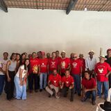 Grupo de pessoas posando em um salão coberto, diante de uma parede clara. Parte do grupo veste camisetas vermelhas com estampas brancas, enquanto outras pessoas usam roupas casuais. Algumas estão em pé e outras agachadas à frente. O espaço tem piso de cerâmica, cadeiras plásticas nas laterais e telhado aparente.