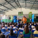 Fotografia de uma apresentação educativa em um espaço coberto, com várias crianças sentadas em cadeiras azuis, de costas para a câmera. À frente, uma pessoa fala ao microfone ao lado de dois personagens fantasiados, um azul e outro laranja. O ambiente tem banners da Saneago, cartazes sobre água e saneamento, caixas de som e estrutura metálica no teto.