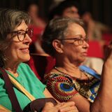 Fotografia de duas mulheres sentadas em uma plateia com poltronas vermelhas. A mulher à esquerda usa blusa verde e óculos, sorrindo enquanto assiste à atividade. A mulher à direita usa óculos, blusa estampada e segura um celular, aparentemente registrando a cena. Ao fundo, outras pessoas aparecem desfocadas na plateia.