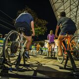 Fotografia noturna de pessoas pedalando em bicicletas ergométricas instaladas em uma área externa coberta. Em primeiro plano, aparecem duas bicicletas vistas por trás, com participantes pedalando. Ao fundo, há pessoas sentadas em cadeiras, iluminação urbana e uma estrutura coberta, sugerindo uma atividade coletiva ao ar livre.