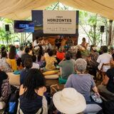Registro de um painel ou roda de conversa realizado sob uma tenda, em ambiente externo e arborizado. Ao centro, há pessoas sentadas em cadeiras participando da discussão, com microfones e estrutura de som. O público acompanha a atividade sentado em cadeiras, almofadas e bancos, disposto em semicírculo. Ao fundo, aparece um painel com a identificação “Fórum Horizontes — Diálogos para novos caminhos” e uma tela lateral com a marca do FICA 2026.