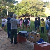 Grupo de crianças e adultos reunidos ao ar livre, em uma área com árvores, gramado e calçada de pedra. Algumas pessoas usam camisetas com identificação institucional, e há caixas e equipamentos apoiados sobre bancos próximos ao grupo. A cena mostra os participantes em roda, aparentemente acompanhando uma explicação ou atividade.