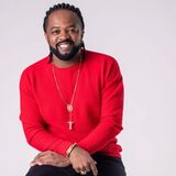 Retrato em estúdio de um homem sorrindo, sentado, usando suéter vermelho, calça escura e acessórios dourados, como colar com pingente de cruz, pulseira e anéis. O fundo é claro e neutro, com iluminação suave.