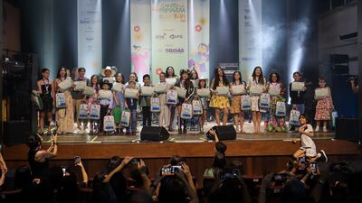 Grupo de crianças e jovens em um palco, segurando certificados e mochilas. Ao fundo, há banners com o texto “4º Canto da Primavera Kids 2025”, ilustrações coloridas e logotipos de instituições. Na plateia, várias pessoas aparecem com celulares levantados, registrando o momento. O palco tem caixas de som, iluminação e fumaça cênica.