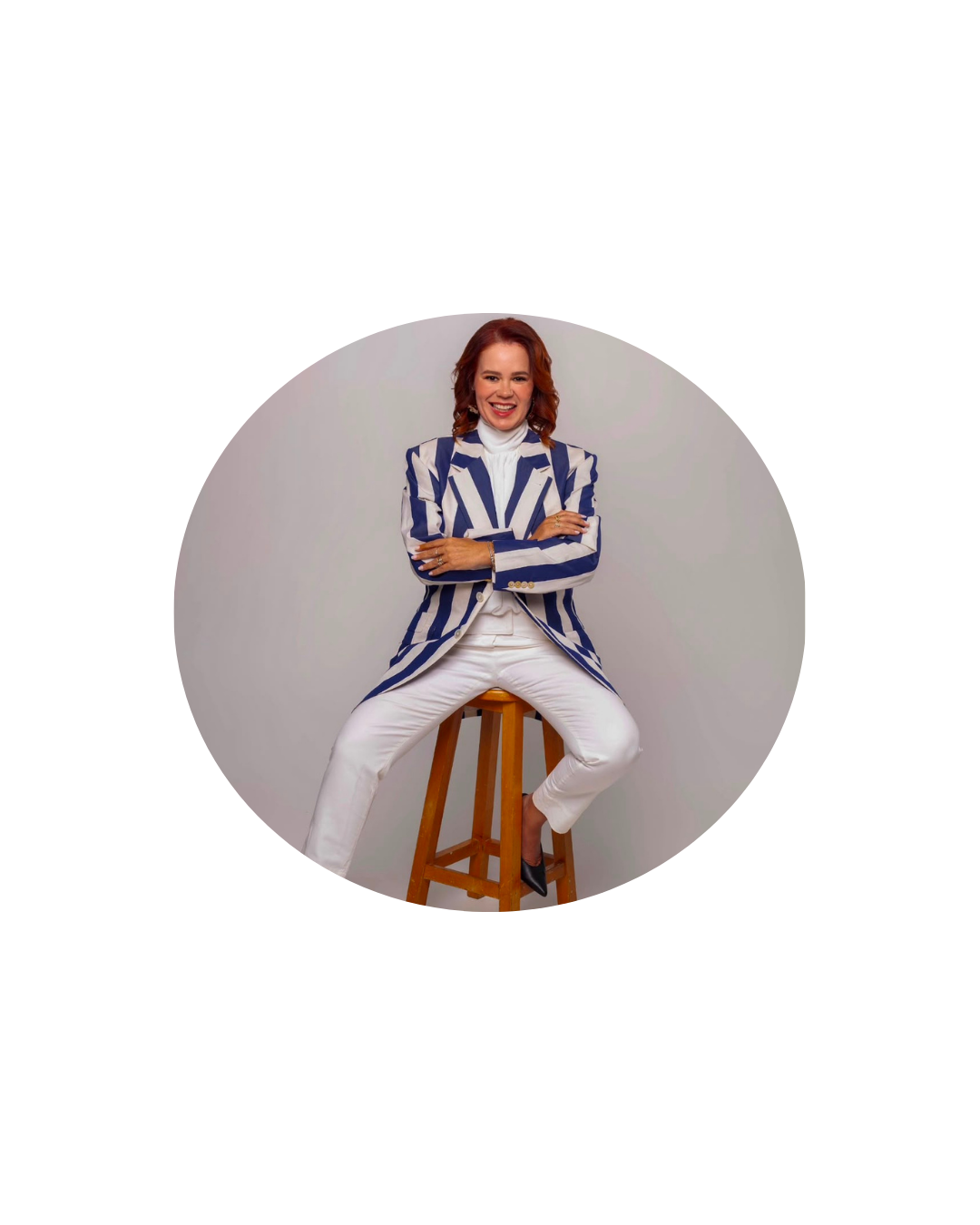 Mulher ruiva sentada em um banco alto de madeira, em pose descontraída e sorrindo diretamente para a câmera. Veste um conjunto composto por calça e blusa brancas, complementado por um blazer branco com listras azuis, além de sapatos escuros. A fotografia foi realizada em estúdio, com fundo neutro e iluminação suave, transmitindo uma imagem profissional e acolhedora.