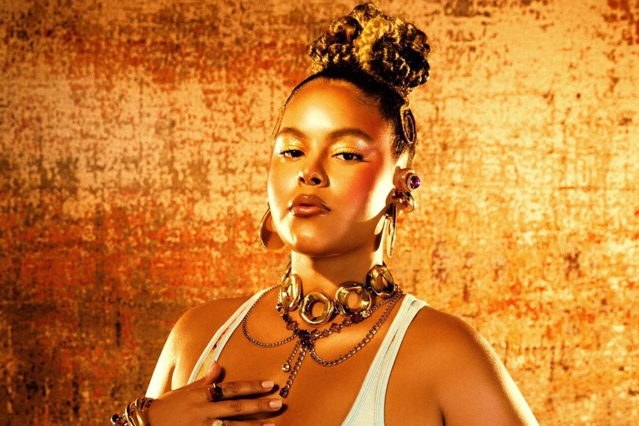 Retrato de uma mulher em pose frontal, enquadrada do busto para cima, diante de um fundo em tons quentes de laranja e dourado com textura. Ela tem pele morena clara, cabelos presos em um coque alto com tranças nas laterais e expressão séria, olhando diretamente para a câmera. Usa uma blusa branca de alças, grandes brincos e um conjunto de colares metálicos dourados em camadas, além de pulseiras e anéis. Uma das mãos repousa sobre o peito. A iluminação quente destaca o rosto, a maquiagem com sombras douradas e o brilho dos acessórios, criando uma composição elegante e marcante.