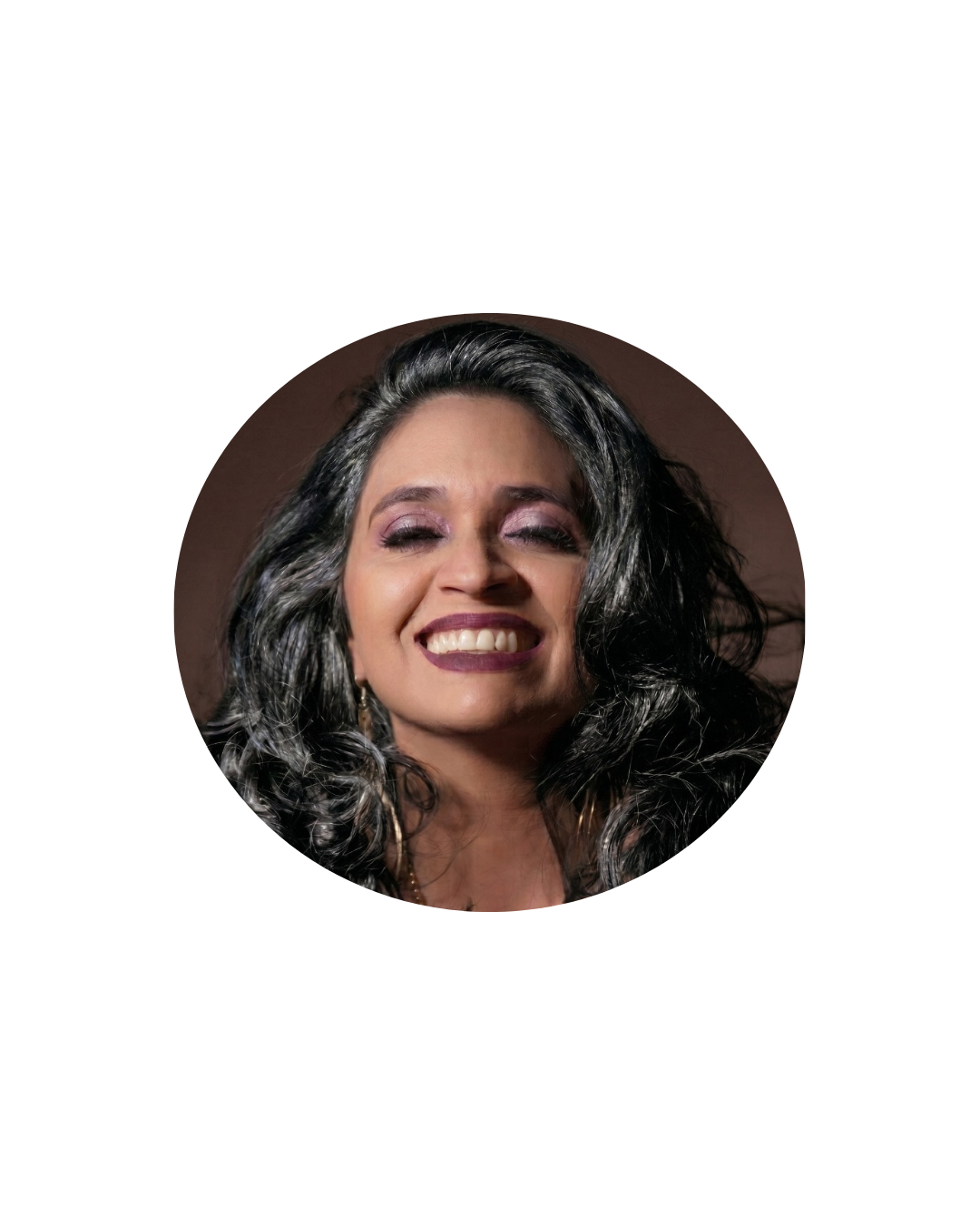 Mulher de cabelos escuros com mechas grisalhas em destaque, registrada em um retrato aproximado. Ela sorri com os olhos fechados, transmitindo espontaneidade e leveza. Usa maquiagem em tons de roxo, batom escuro e brincos longos, enquanto o fundo desfocado valoriza sua expressão facial e confere um aspecto artístico à composição.
