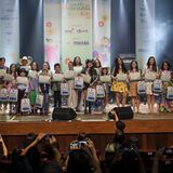 Grupo de crianças e jovens em um palco, segurando certificados e mochilas. Ao fundo, há banners com o texto “4º Canto da Primavera Kids 2025”, ilustrações coloridas e logotipos de instituições. Na plateia, várias pessoas aparecem com celulares levantados, registrando o momento. O palco tem caixas de som, iluminação e fumaça cênica.