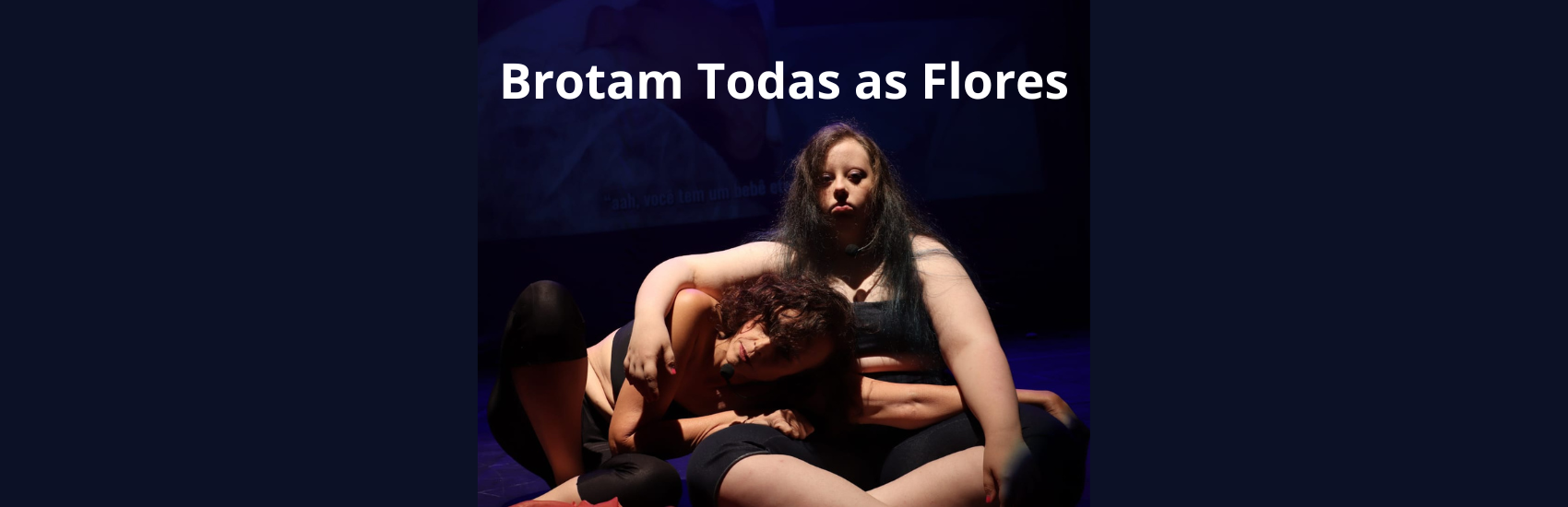 Duas mulheres em um foco de luz. Uma mulher jovem, branca, com cabelos longos e com síndrome de Down, sentada com pernas cruzadas, olha para frente séria. Abraça outra mulher de meia idade, branca, cabelos na altura dos ombros,  que deita no seu colo. As duas usam calça e top pretos em fundo escuro.