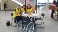 Foto estão três colaboradores do Núcleo de Acessibilidade, sentados e sorrindo, da direita para a esquerda Leandro Viana e Dhenny Ketully (intérpretes de Libras) e Juliana Barbosa (secretária do Núcleo). Todos estão sentados atrás de uma mesa.