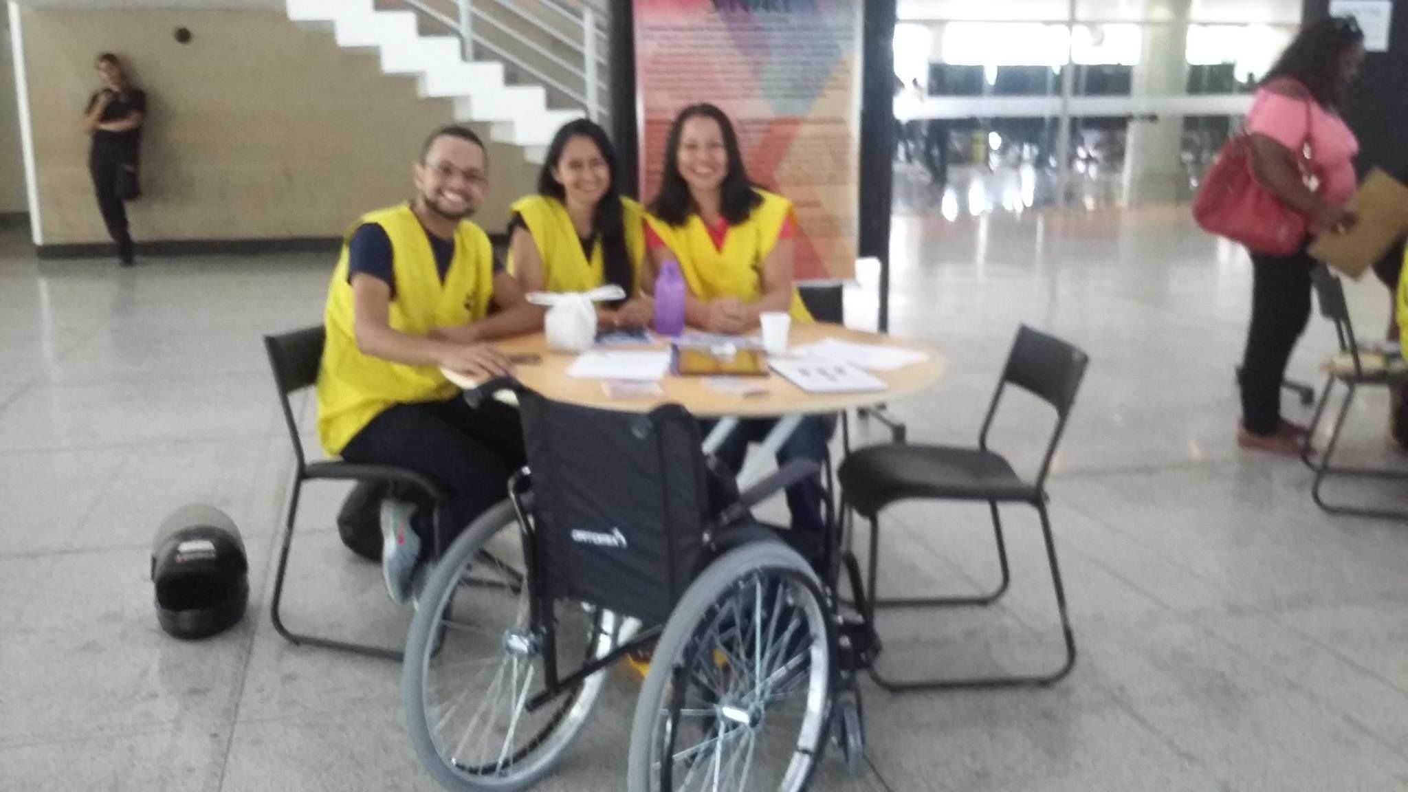 Na foto estão três colaboradores do Núcleo de Acessibilidade, sentados e sorrindo, da direita para a esquerda Leandro Viana e Dhenny Ketully (intérpretes de Libras) e  Juliana Barbosa (secretária do Núcleo). Todos estão sentados atrás de uma mesa.